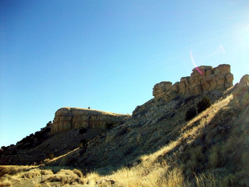 A panoramic view of rocky hills under a clear blue sky, with sunlight highlighting the textured formations and golden grasses in the foreground. South Shore Lake Pueblo mountain bike trail.