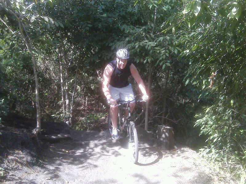 A person riding a mountain bike on a narrow trail surrounded by dense greenery. The cyclist is wearing a helmet and athletic clothing, navigating through a natural setting. Markham Park mountain bike trail.