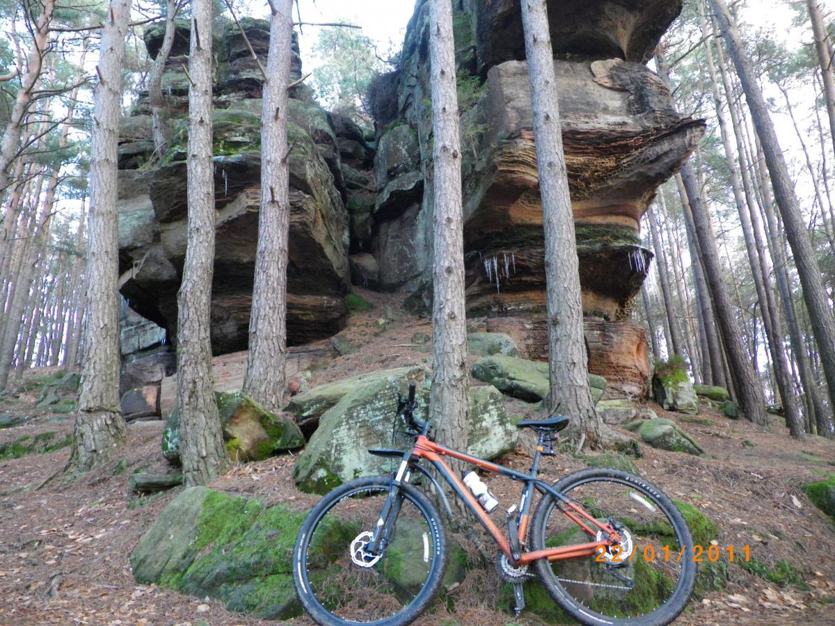 A mountain bike parked on the forest floor next to large rock formations and tall pine trees. The scene is natural and tranquil, with moss-covered rocks and some icicles hanging from the rocks above. Rodalben F Trail mountain bike trail.