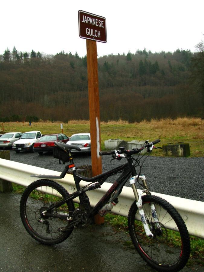 A mountain bike parked by a roadside sign that says "Japanese Gulch." In the background, there is a grassy area with trees, and several parked cars are visible nearby. The sky is overcast, suggesting a rainy day. Japanese Gulch mountain bike trail.