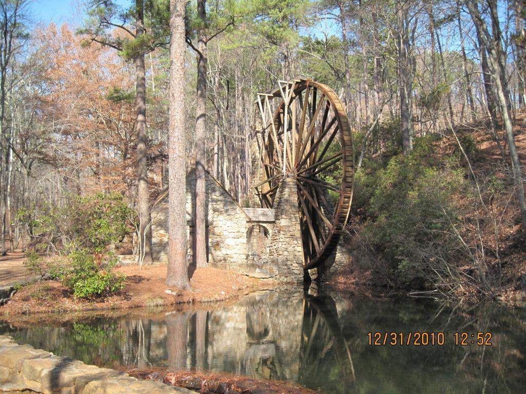 An old wooden water wheel beside a stone structure, partially surrounded by trees and reflected in a calm pond. The scene captures a serene natural setting, with a mix of evergreen and deciduous trees in the background. Berry College mountain bike trail.