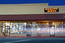 A storefront with a sign that reads "REALITY BIKES," featuring large windows displaying bicycle-related items. The building is illuminated, set against a twilight sky, with light trails visible, suggesting motion outside.