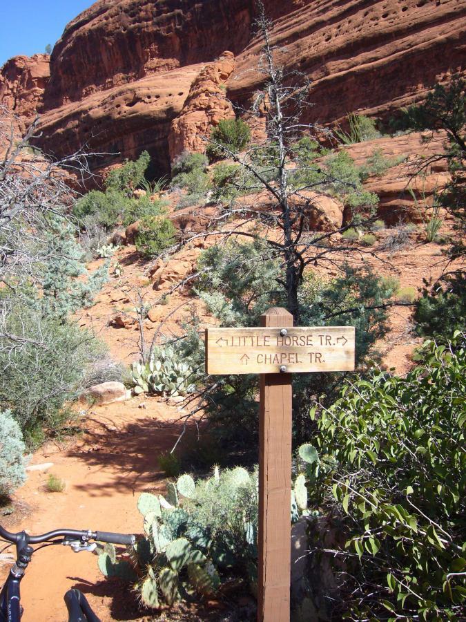 A wooden trail sign indicating directions to "Little Horse Trail" and "Chapel Trail," set against a backdrop of red rock formations and desert vegetation. Bell Rock Trailway mountain bike trail.
