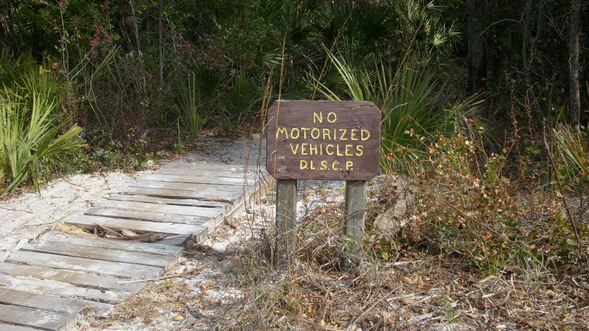 A wooden sign stating "No Motorized Vehicles" in yellow letters, positioned near a sandy path bordered by greenery and small plants. The path is made of wooden planks and leads into a dense wooded area. Spruce Creek Preserve mountain bike trail.