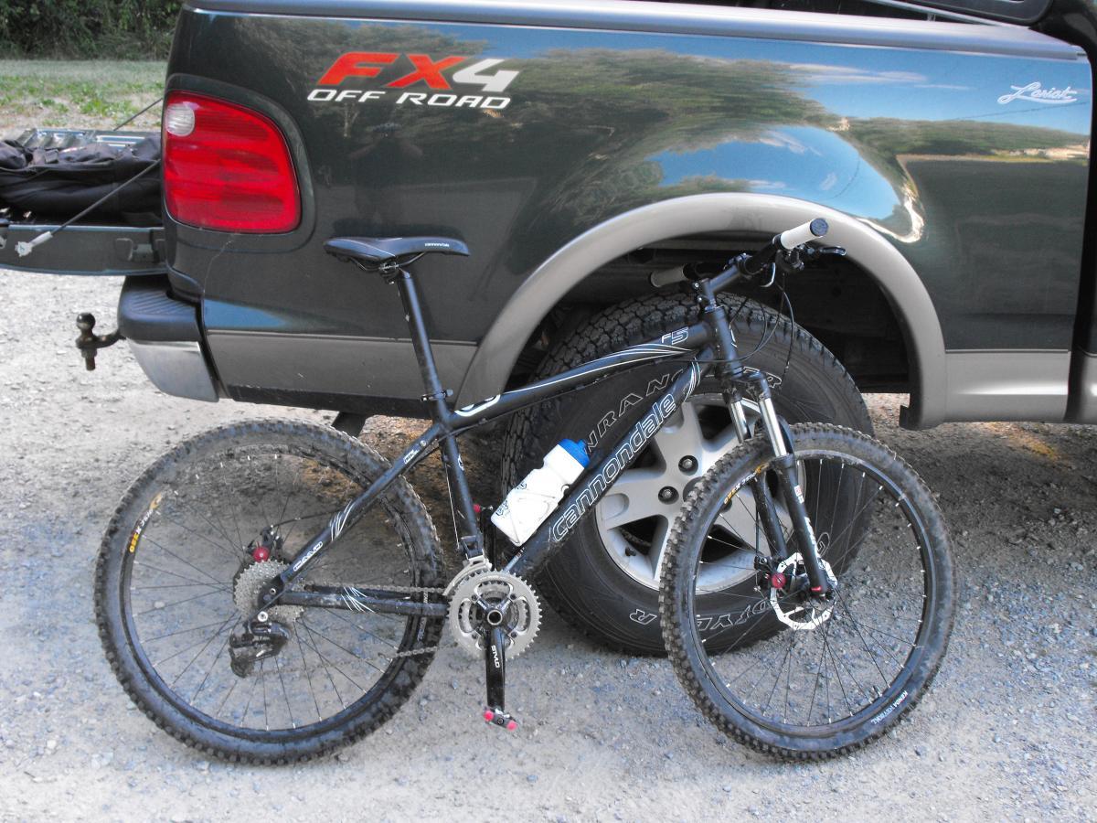 Alt tag: A black Cannondale mountain bike with a water bottle mounted on the frame, leaning against the rear tire of a green pickup truck, parked on a gravel path. Greenbrier State Park mountain bike trail.