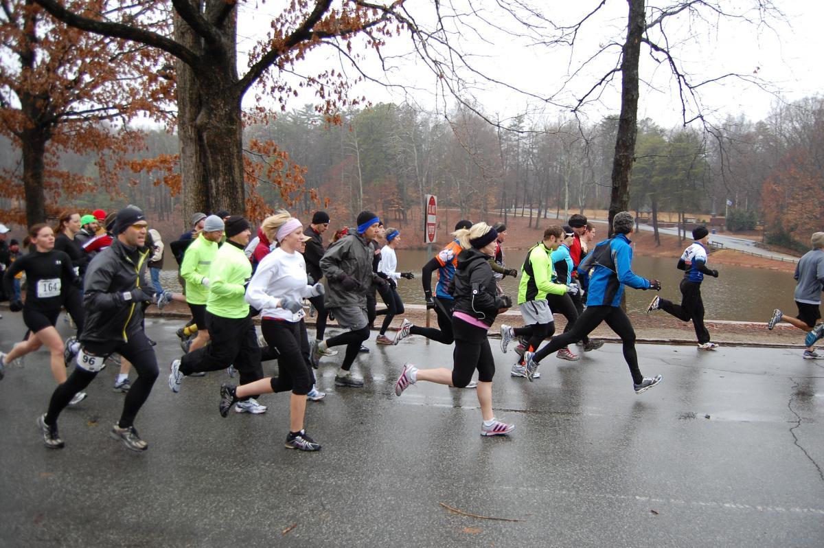 A group of runners participating in a race during rainy weather, with a park setting featuring bare trees and a pond in the background. Some runners are wearing brightly colored athletic gear, while others are dressed in darker clothing. Country Park mountain bike trail.