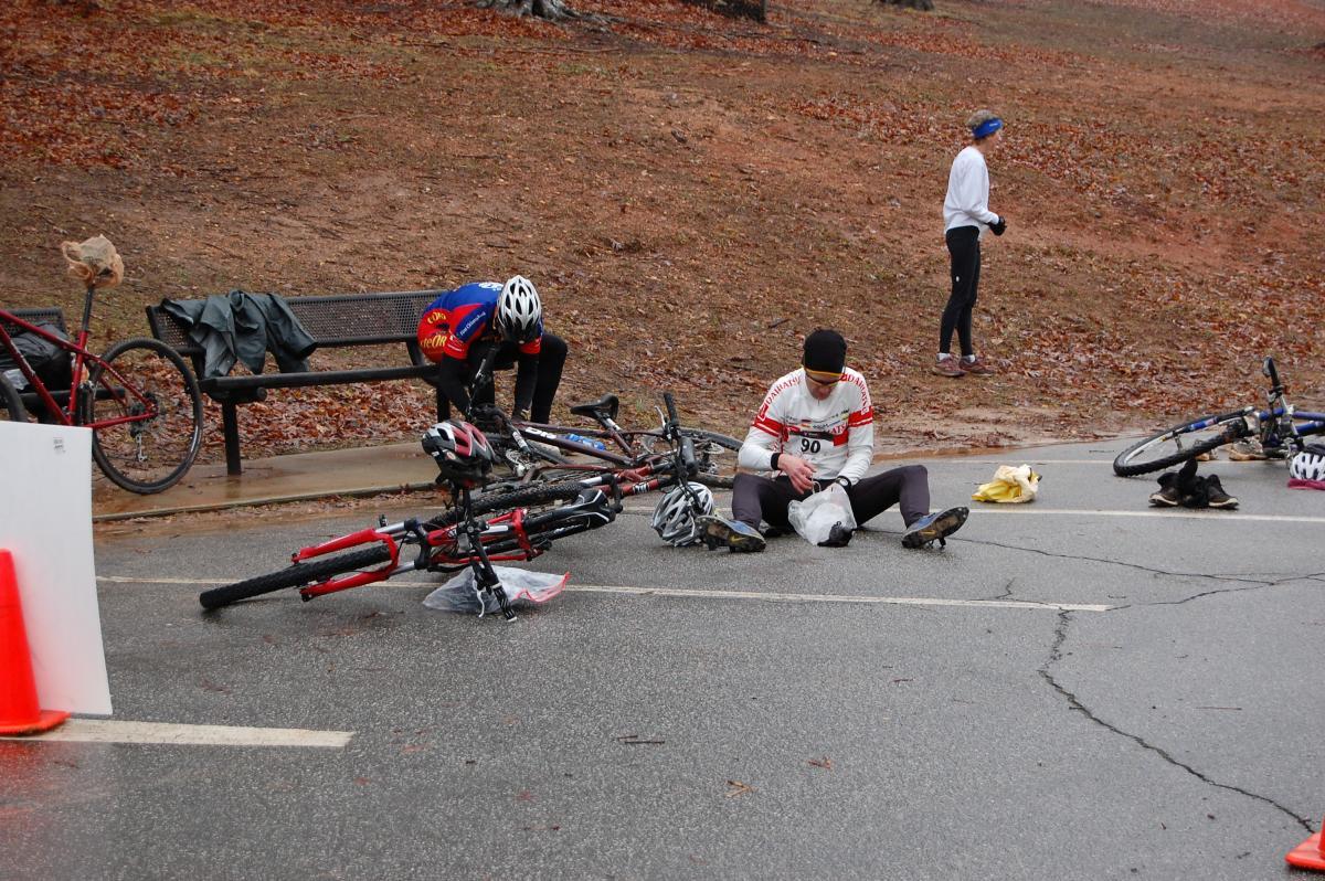Two cyclists are seen on the ground near their bicycles, which are lying on the road. One cyclist is sitting and inspecting an item while the second is hunched over their bike. A bench and a person in the background are also visible, with fallen leaves scattered across the ground, indicating a cloudy or overcast day. A traffic cone is positioned in the foreground. Country Park mountain bike trail.