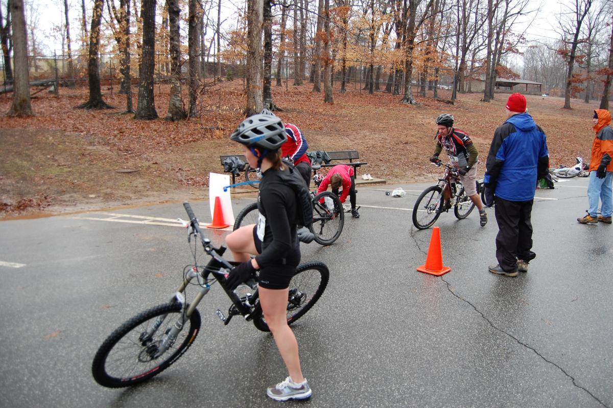 A group of cyclists in a wooded area during a rainy event. One cyclist, wearing a black outfit and helmet, rides a mountain bike past orange cones, while another cyclist adjusts their bike on the ground. Two other participants are engaged in preparation, and spectators in rain gear are visible nearby. The ground is wet, and the trees in the background are bare, indicating a cool season. Country Park mountain bike trail.