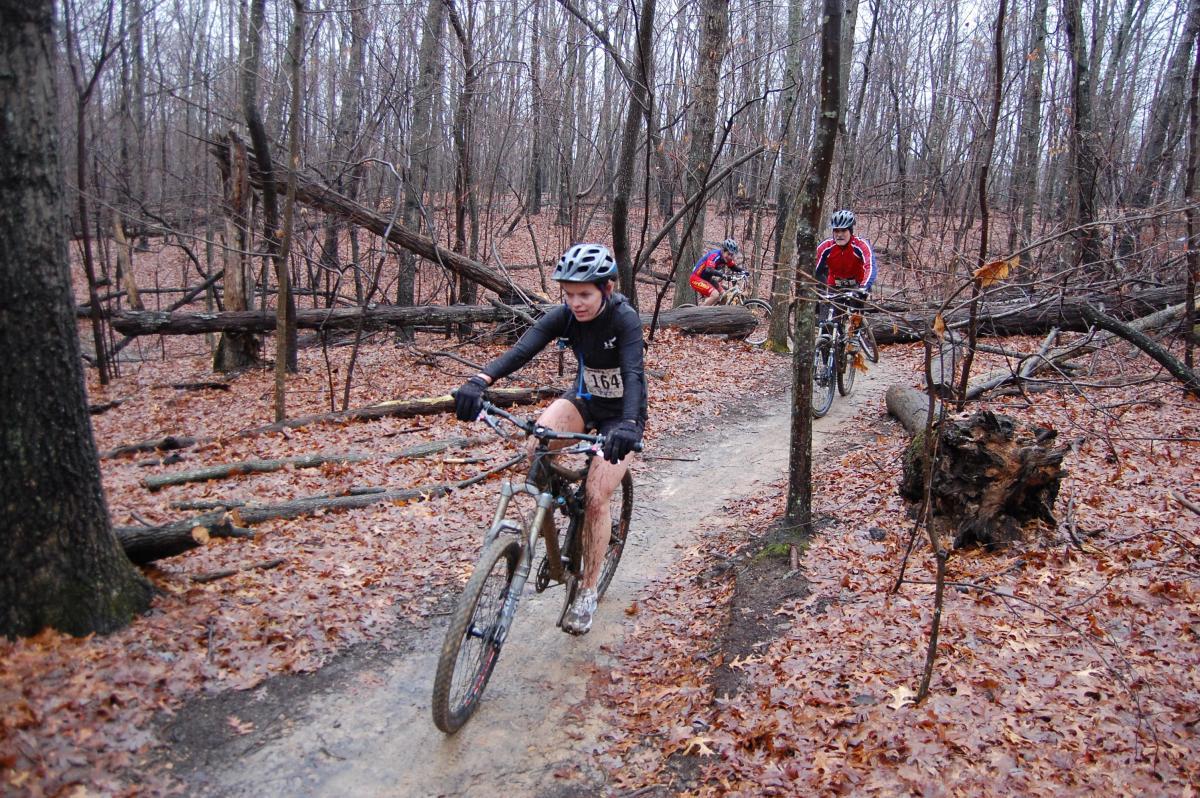 A group of mountain bikers navigating a dirt trail through a dense forest in autumn. The ground is covered with brown leaves, and there are fallen trees along the path. One cyclist is in the foreground wearing a black outfit and helmet, while two others are visible in the background. The atmosphere appears calm and slightly overcast. Country Park mountain bike trail.