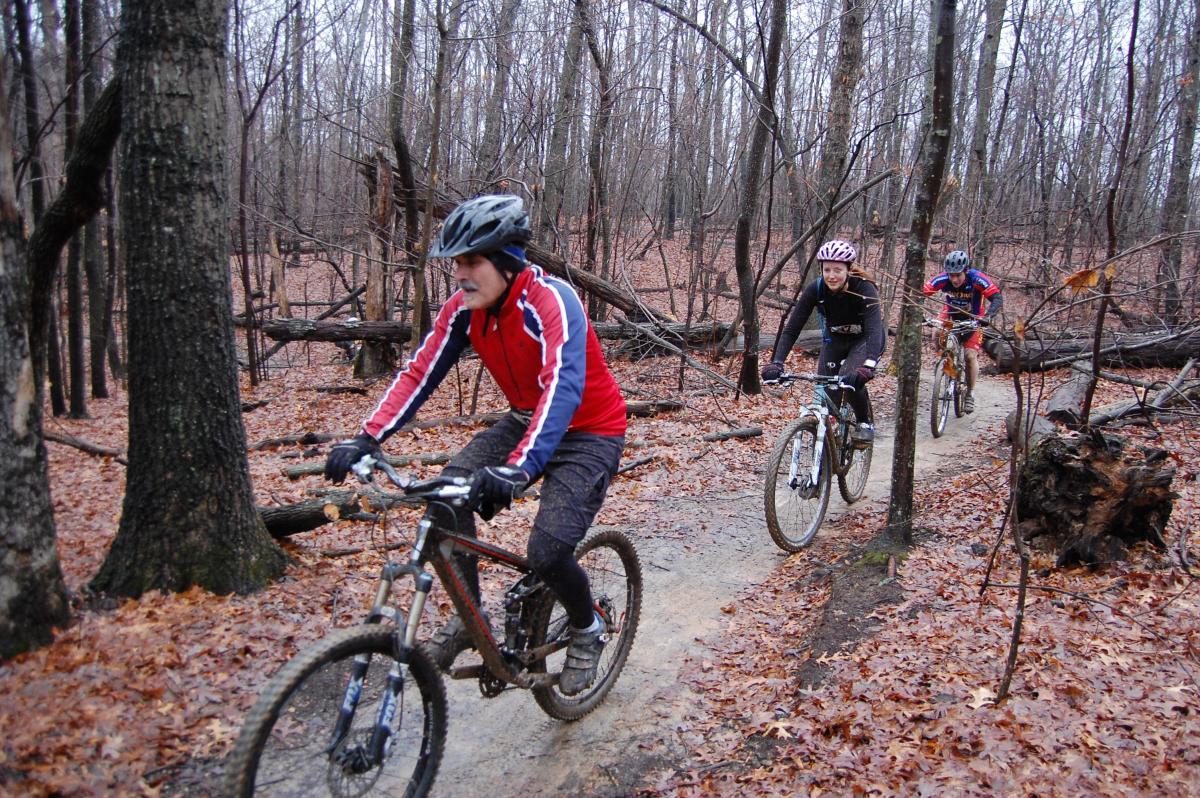 Three mountain bikers navigate a dirt trail through a wooded area with bare trees in early spring. The first rider, wearing a red and blue jacket, is leading the group, while the second rider, a woman in a black outfit and helmet, smiles as she bikes closely behind. The third rider, in a blue and red jersey, trails behind them. The ground is covered with fallen leaves, and some tree branches lay scattered along the path. Country Park mountain bike trail.
