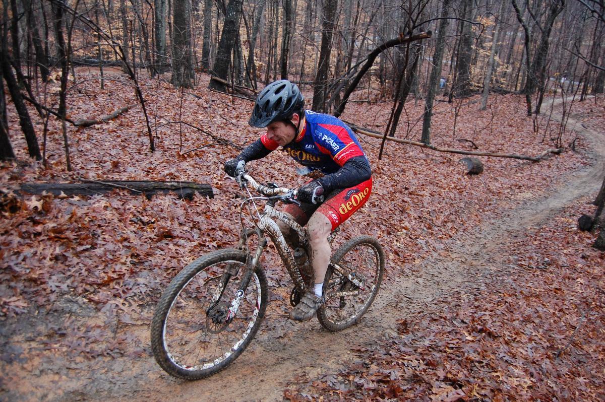 A mountain biker navigating a muddy trail covered with fallen leaves, dressed in a blue and red cycling jersey and black shorts, focused intently as he climbs uphill, with trees in the background. Country Park mountain bike trail.