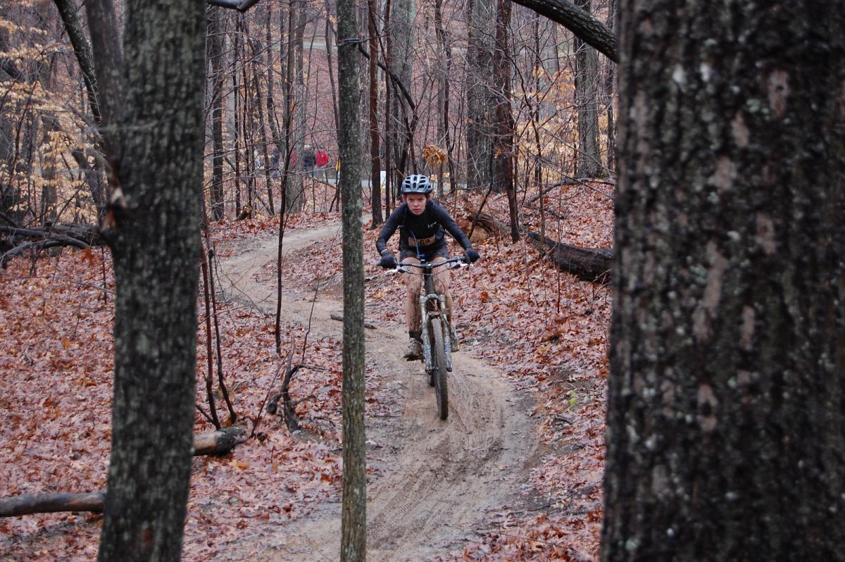 A mountain biker navigating a dirt trail in a forest during autumn, surrounded by trees with fallen leaves. The rider is focused as they traverse the winding path, showcasing the dynamic nature of the sport amidst a natural setting. Country Park mountain bike trail.