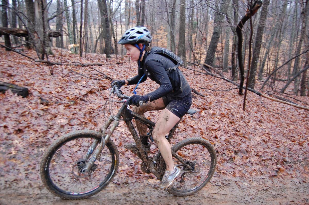 A mountain biker navigating a muddy trail, wearing a helmet and athletic gear. The biker's legs and bike are splattered with mud, surrounded by a forest of trees and fallen leaves. Country Park mountain bike trail.