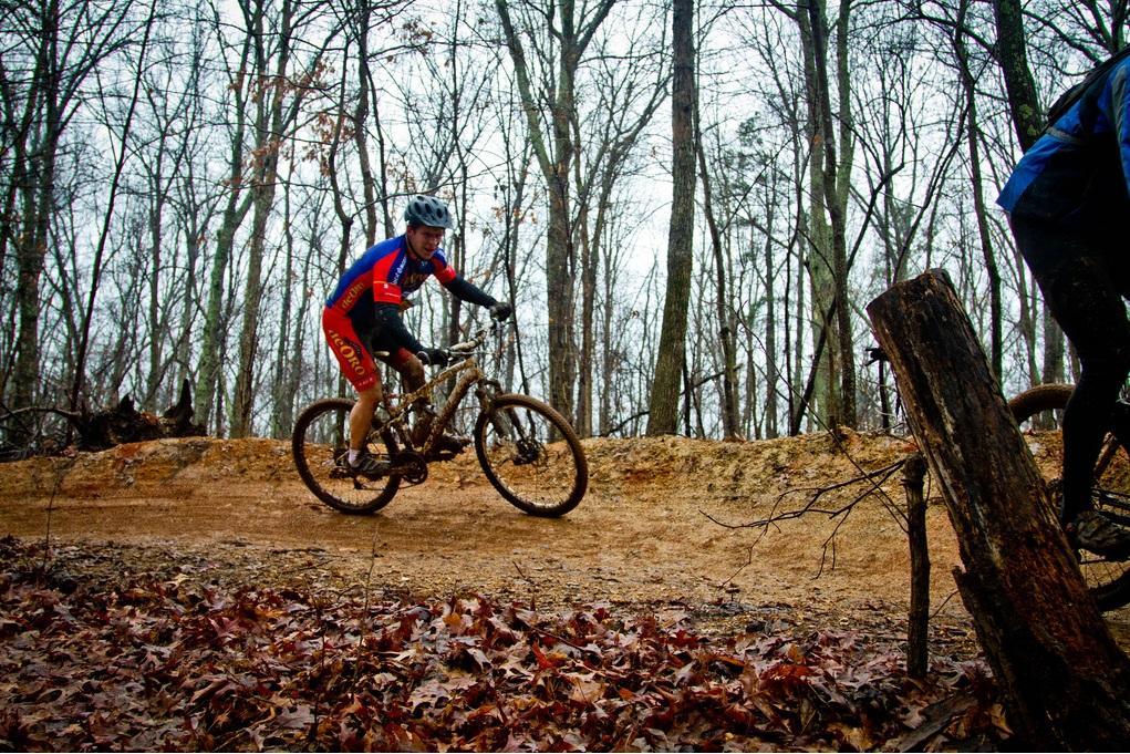 A mountain biker dressed in a red and blue jersey rides along a dirt trail in a wooded area during an overcast day. The scene features sparse trees and fallen leaves scattered on the ground, capturing the essence of outdoor cycling in nature. Country Park mountain bike trail.