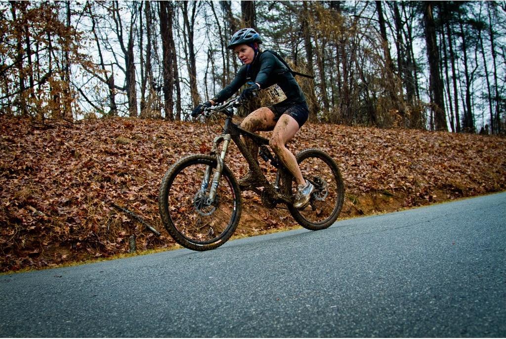A cyclist riding a mountain bike along a roadway, covered in mud, with autumn leaves scattered on the ground and trees in the background. The cyclist is wearing a helmet and athletic attire, showcasing the physicality of the sport in a wooded environment. Country Park mountain bike trail.