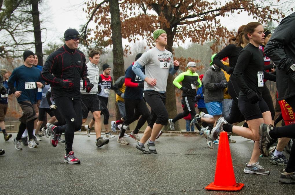 A group of runners in athletic gear participate in a race on a rainy day. The scene includes various individuals wearing numbered bibs as they run on a wet road, with trees in the background. The atmosphere captures the energy of a competitive outdoor event despite the inclement weather. Country Park mountain bike trail.