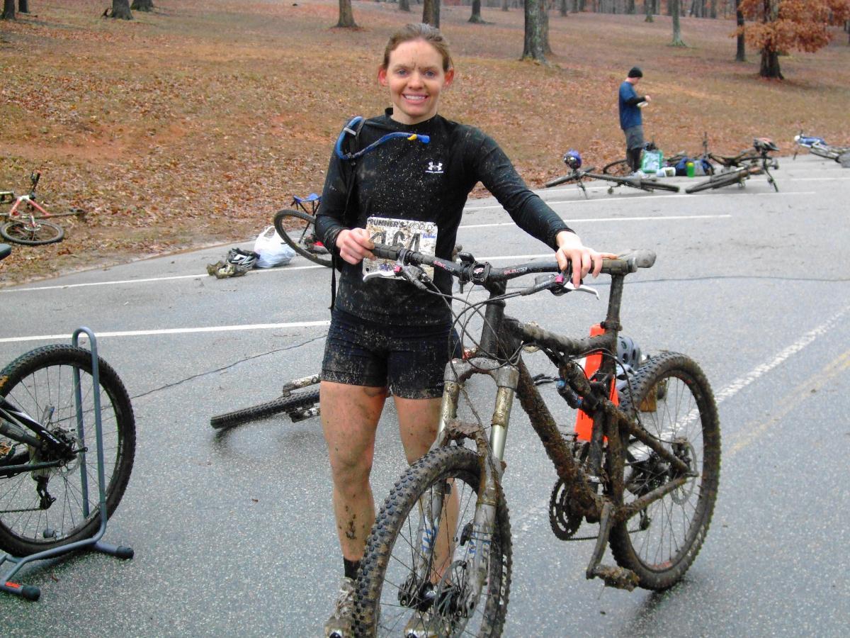 A smiling cyclist covered in mud stands beside a mountain bike in a forested area. The cyclist is wearing a black athletic outfit and has a race number pinned to their chest. In the background, other bikes and individuals are visible, suggesting a recent biking event. The ground is littered with fallen leaves, indicating an outdoor setting. Country Park mountain bike trail.
