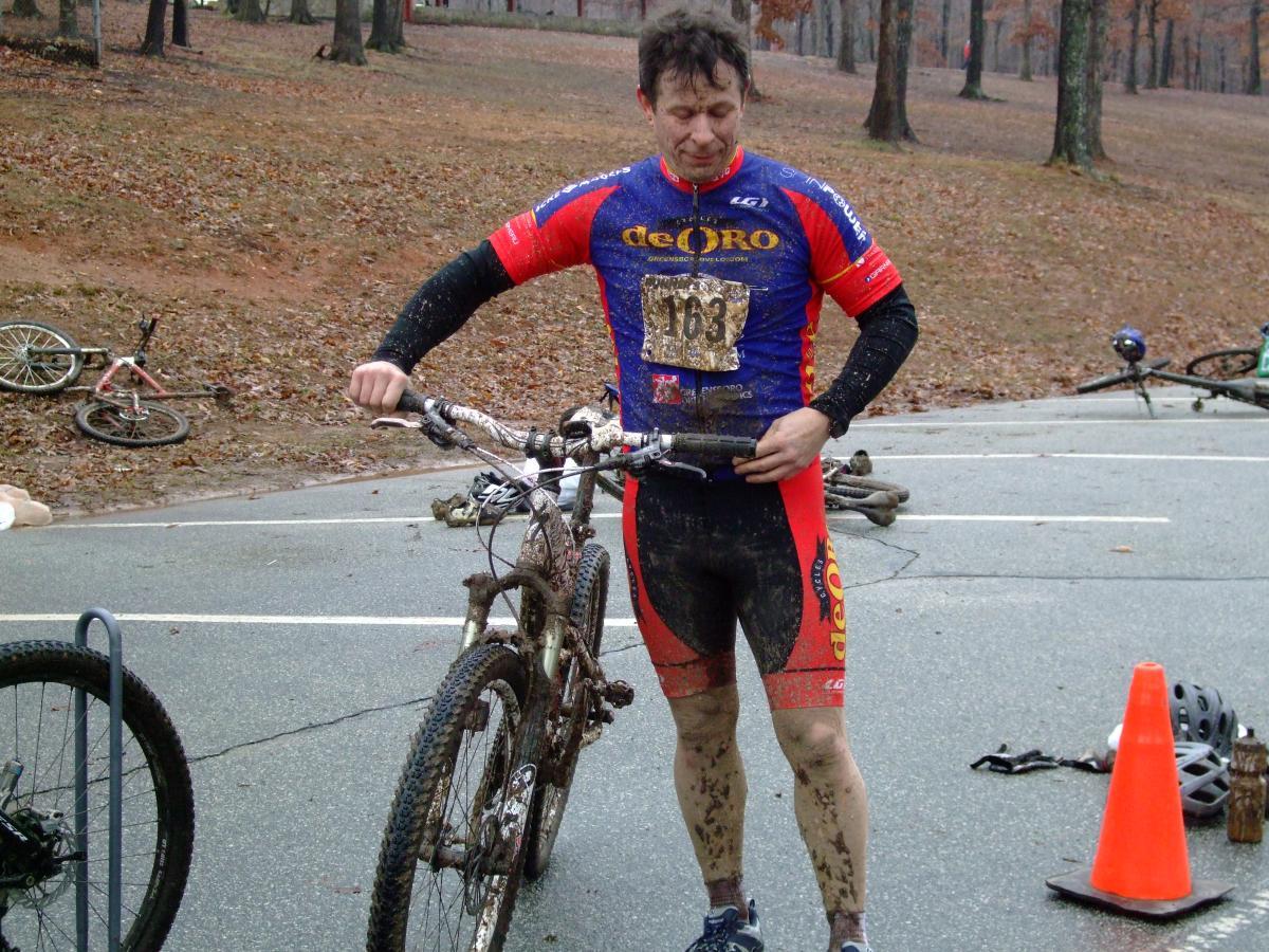 A mountain biker in a blue and red racing jersey stands beside his bike, which is covered in mud. He appears slightly dirty and is inspecting the bike in a park setting with trees in the background. A few bicycles can be seen in the background, along with a traffic cone on the ground. Country Park mountain bike trail.