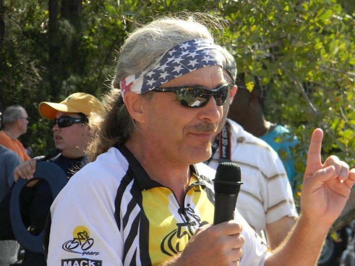 A man with long, gray hair and a patriotic headband speaks into a microphone, gesturing with his right hand. He is wearing sunglasses and a cycling jersey. In the background, a small crowd is visible, including a person wearing an orange cap. The setting appears outdoor, surrounded by greenery. Virginia Key North Point mountain bike trail.