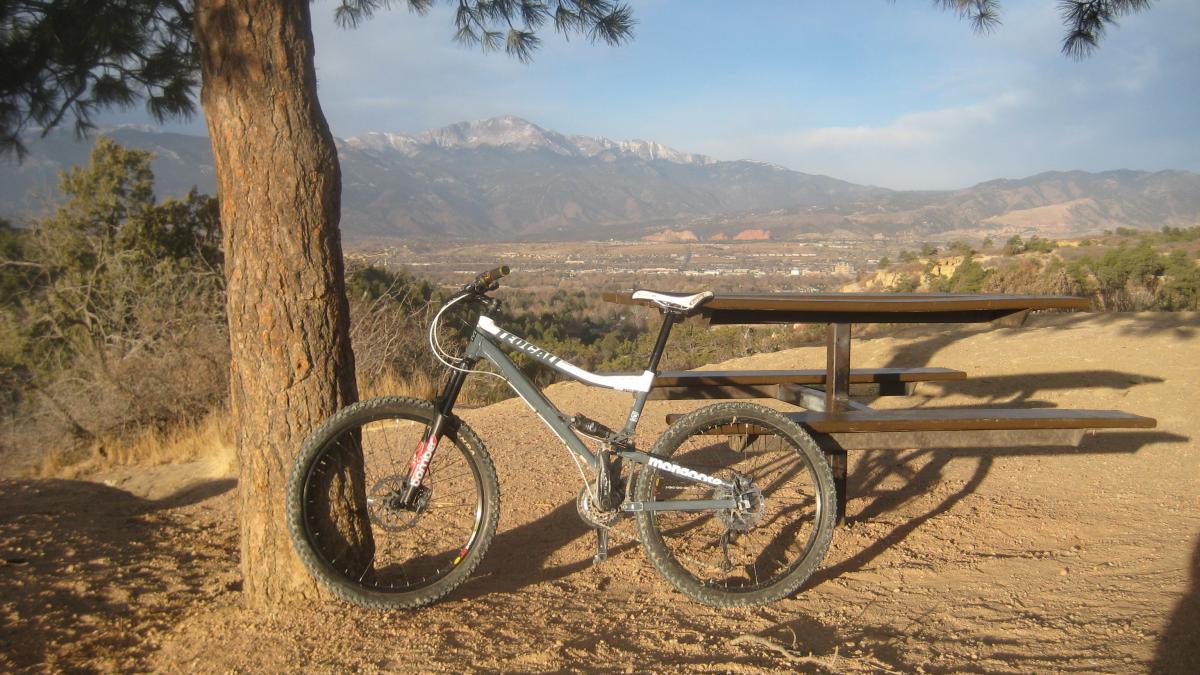 A mountain bike leaning against a tree in a scenic outdoor setting, with a picnic table in the background and mountains visible in the distance under a partly cloudy sky. Palmer Park mountain bike trail.