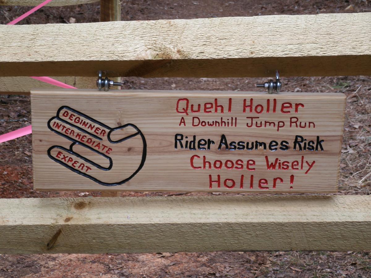A wooden sign hanging on a fence with text indicating the "Quehl Holler" downhill jump run. The sign features a hand diagram with labels for "Beginner," "Intermediate," and "Expert" levels, along with cautionary phrases like "Rider Assumes Risk" and "Choose Wisely Holler!" The background shows a wooded area with pink ribbon marking. Blankets Creek mountain bike trail.