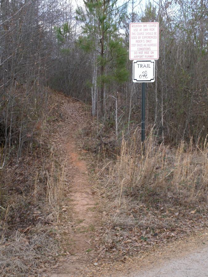 A narrow dirt trail leading into a wooded area, flanked by trees and dry grass. A sign post on the right warns that the mountain bike course is for experienced riders only and includes instructions about safety and restrictions on riding near the airport runway. Kiwanis Fairground mountain bike trail.