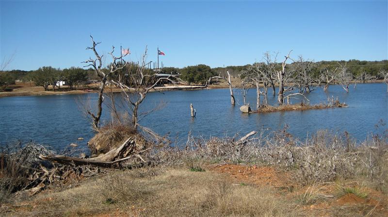 A serene landscape featuring a calm body of water surrounded by dry, leafless trees and grassy shorelines. In the background, there are flags, likely indicating a nearby facility, and rocky outcrops are partially visible in the water. The scene is under a clear blue sky, suggesting a peaceful, sunny day. Reveille Peak Ranch mountain bike trail.