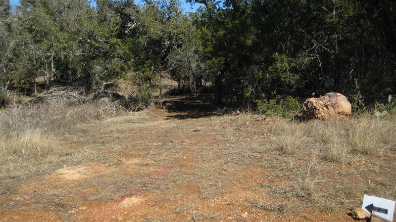 A dirt path leading into a wooded area, surrounded by trees and sparse grasses. In the foreground, a large rock is visible to the right, and a small sign points left off the path. The scene is well-lit with clear blue skies above. Reveille Peak Ranch mountain bike trail.