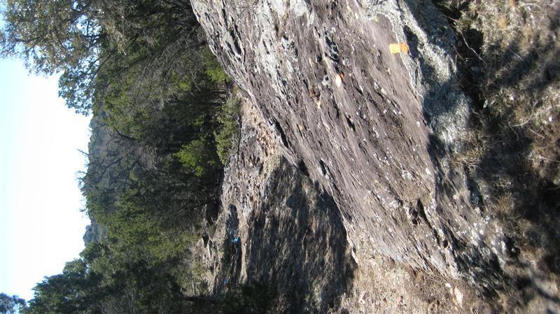Sunlit rocky terrain with rough surfaces and patches of greenery, showing a large rock formation and scattered vegetation in the background. Reveille Peak Ranch mountain bike trail.