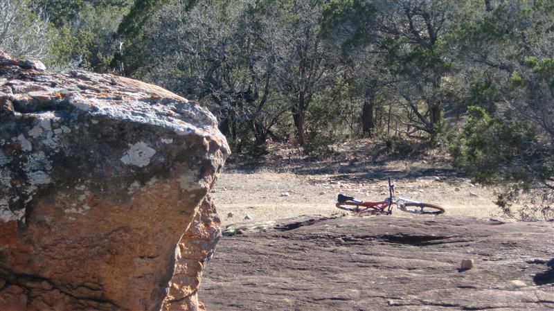 A rocky landscape with a large, textured boulder in the foreground, while a mountain bike is visible in the background, partially obscured by trees and vegetation. Reveille Peak Ranch mountain bike trail.