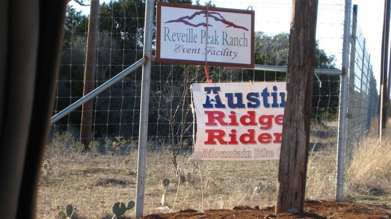 A sign for Revelle Peak Ranch Event Facility is mounted on a fence, accompanied by a banner for Austin Ridge Riders, a mountain bike organization. The scene includes surrounding natural vegetation and a clear sky. Reveille Peak Ranch mountain bike trail.