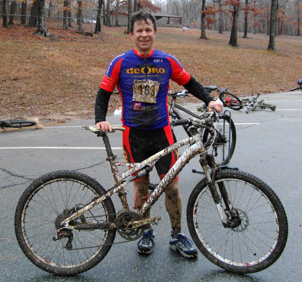 A mud-covered cyclist stands outdoors next to a muddy mountain bike, wearing a blue and red jersey with the number 163. The background features a leaf-covered landscape with trees, and other bikes can be seen scattered on the ground. The cyclist has a cheerful expression, suggesting a successful ride despite the muddy conditions. Country Park mountain bike trail.