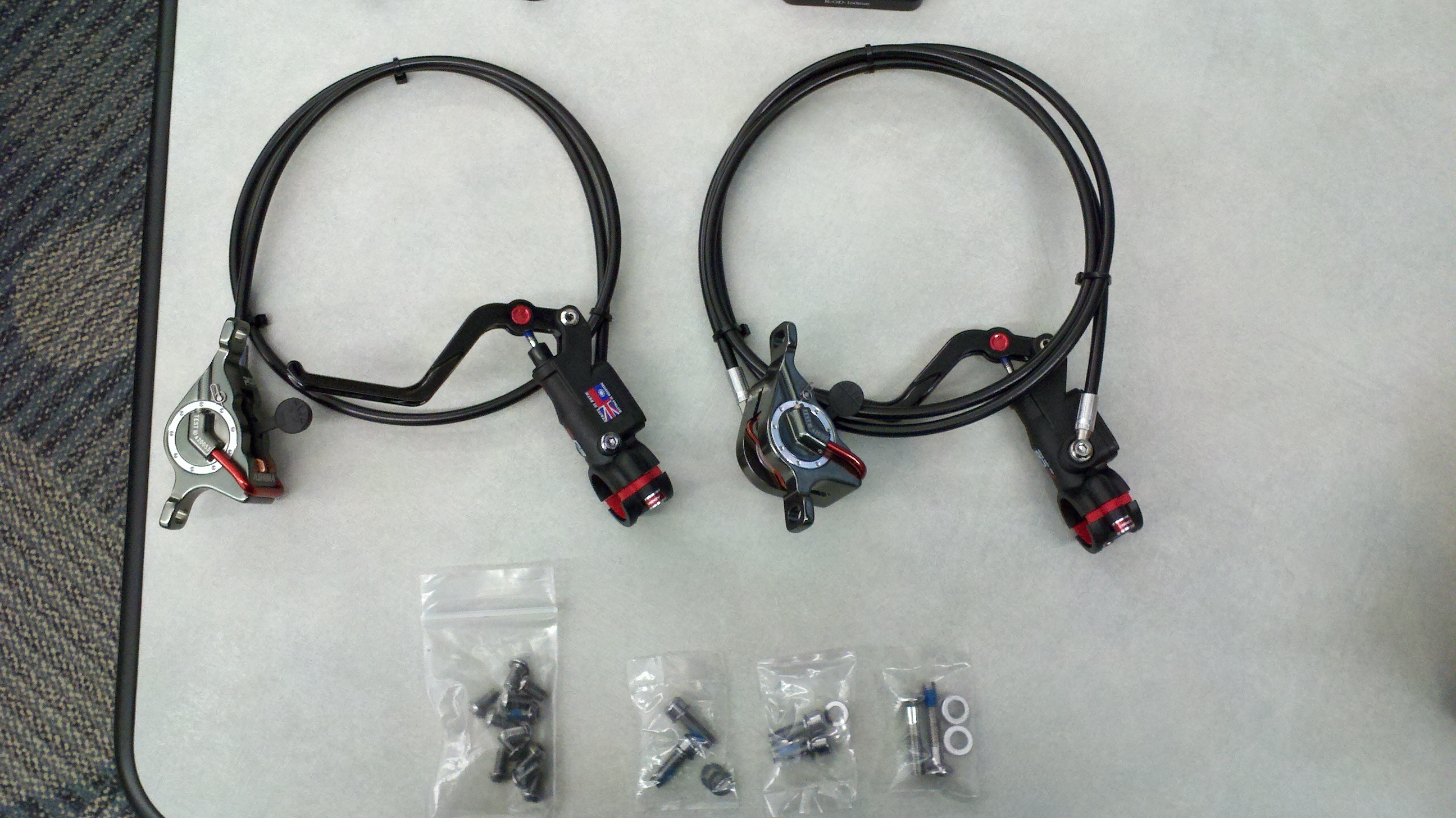 Image of two bike brake levers with cables, arranged side by side on a table, with several small bags of screws and fittings placed underneath. The brake levers are designed for a mechanical braking system.