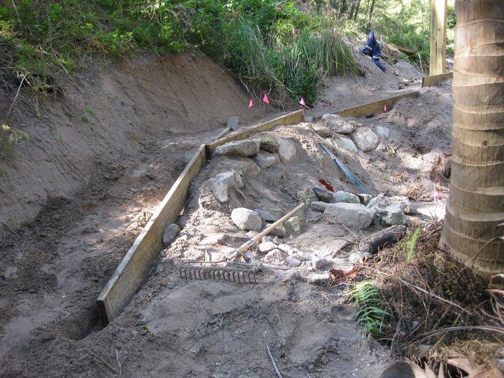 Construction site featuring a partially dug trench with exposed rocks and a wooden frame. Tools like a rake and a blue shovel are visible, along with pink markers indicating the area. Lush greenery surrounds the site, and a palm tree is situated to the right. Virginia Key North Point mountain bike trail.