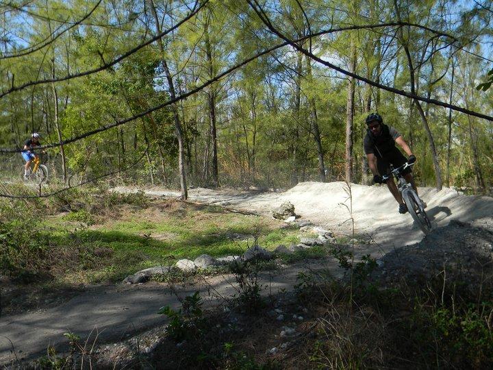 Two mountain bikers navigate a dirt trail surrounded by trees. One rider is in the foreground, leaning into a curve, while the other is seen in the background. The terrain features a mix of dirt and greenery, with sunlight filtering through the foliage. Virginia Key North Point mountain bike trail.