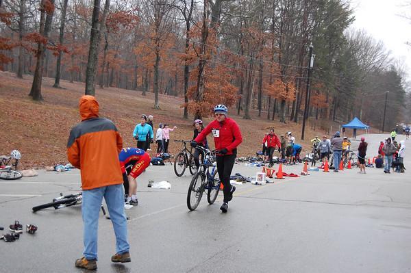 A group of athletes participating in a cycling event in a wooded area. In the foreground, a man in a red shirt is walking with a bike, while another competitor bends down near their bicycle. Several participants are visible in the background, some preparing to ride, amidst scattered gear and cones marking the event area. The trees are bare, indicating a cool season, and a tent is set up in the distance. Country Park mountain bike trail.