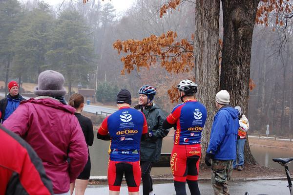 A group of cyclists in colorful jerseys gather by a lake on a cloudy day. They are engaged in conversation, with some wearing jackets and hats to stay warm. Trees with brown leaves and a misty background create a serene atmosphere. Country Park mountain bike trail.