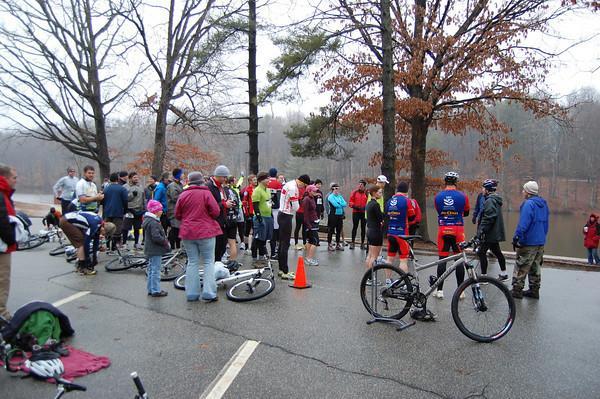 A group of cyclists and spectators gathered on a rainy day in a park. Some participants are on bicycles while others are chatting and preparing for an event. The scene includes trees with sparse leaves and a few orange traffic cones on the ground. Country Park mountain bike trail.