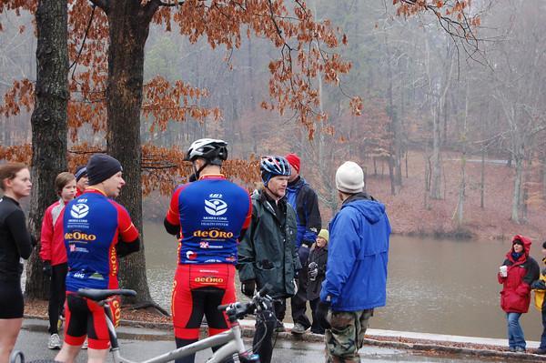 A group of people is gathered near a lake on a cloudy day. Two cyclists in red and blue jerseys are chatting with three others, including a person in a dark jacket, a man in a white hat, and another individual wearing camo pants. In the background, additional spectators are seen watching the scene. The area has barren trees, indicating early winter, and the atmosphere appears casual and social. Country Park mountain bike trail.