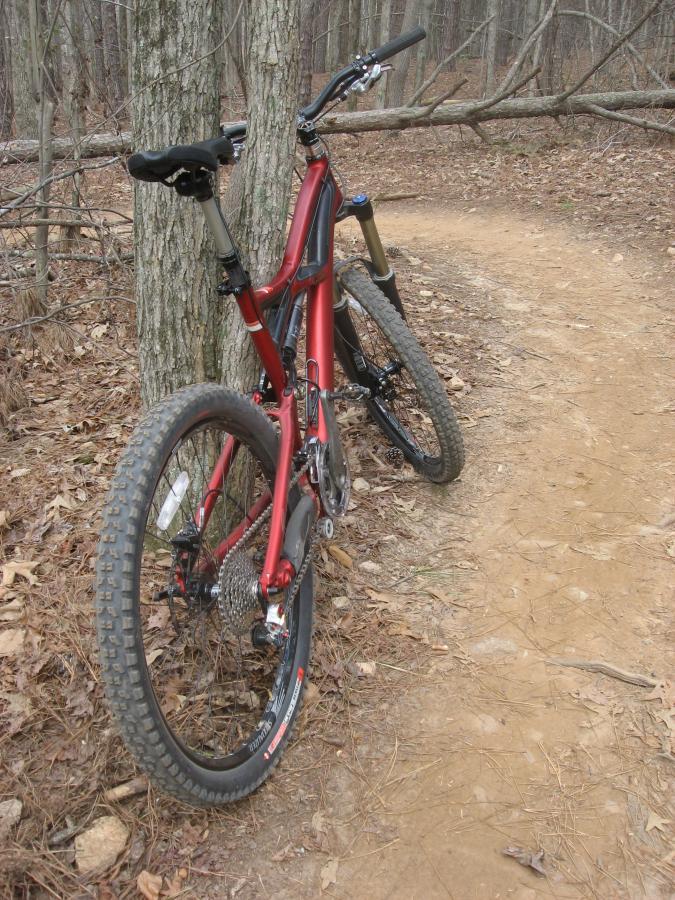 A red mountain bike leaning against a tree on a dirt path in a wooded area, surrounded by fallen leaves and twigs. The trail is narrow and winding, with a natural landscape typical of outdoor biking trails. Big Creek mountain bike trail.