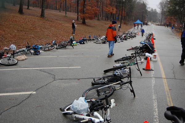 A row of bicycles lying on the ground along a deserted road, with a few people walking nearby. Some bicycles are scattered with gear and supplies around them, and orange traffic cones are placed on one side. In the background, trees with autumn leaves and a blue tent are visible. Country Park mountain bike trail.