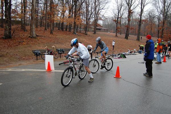 A group of athletes participating in a triathlon, riding mountain bikes along a rain-slicked road lined with orange cones. In the background, other competitors prepare with their bikes and gear, while trees with few leaves indicate a cool, possibly autumn season. Country Park mountain bike trail.