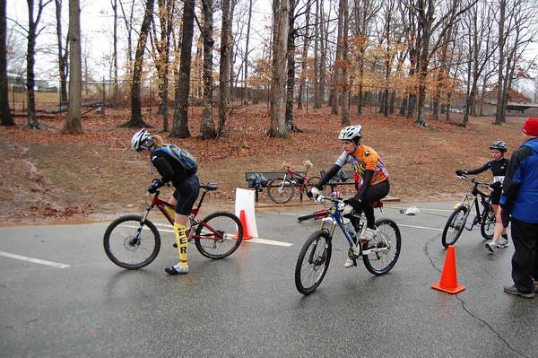 A group of cyclists in athletic gear maneuvering their mountain bikes on a wet road during a race. The scene is set among bare trees, indicating a cool season. There are orange traffic cones marking the course, and some bicycles are propped up nearby. The focus is on two cyclists, one adjusting their bike while another prepares to continue riding. Country Park mountain bike trail.