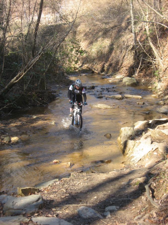 A mountain biker riding through a shallow, rocky creek surrounded by trees and dry foliage, with splashes of water visible as the bike traverses the water. The scene captures the adventurous spirit of outdoor cycling in a natural landscape. Chicopee Woods mountain bike trail.