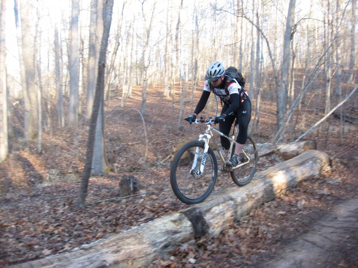 A cyclist navigates a forest trail, skillfully balancing on a fallen log among trees with bare branches and a carpet of dried leaves. The rider wears a helmet and cycling gear, showcasing an active mountain biking scene in a natural setting. Chicopee Woods mountain bike trail.