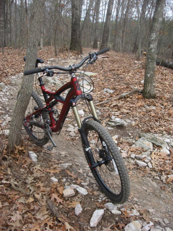 A mountain bike with a red frame is leaning against a tree along a dirt trail covered with rocks and dry leaves. The background features a wooded area with trees and a natural setting, suggesting an outdoor biking scene. Big Creek mountain bike trail.