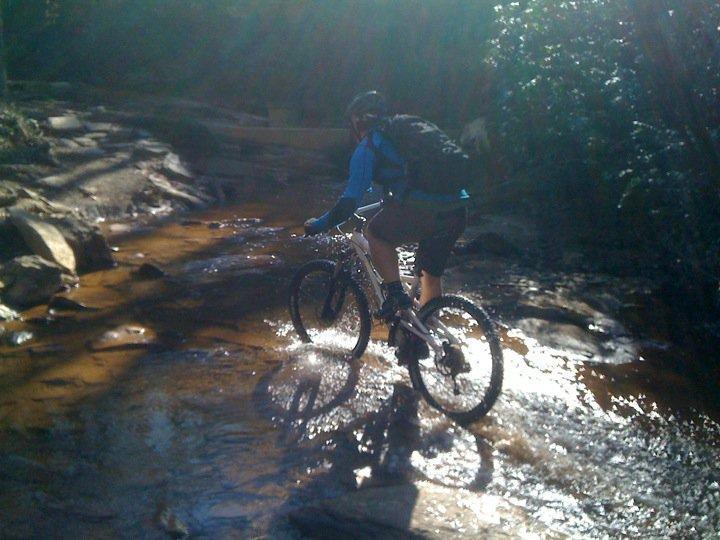 A mountain biker riding through a shallow stream on a rocky trail, with sunlight filtering through the trees above. The rider wears a helmet and a blue shirt, navigating the water and terrain. Chicopee Woods mountain bike trail.
