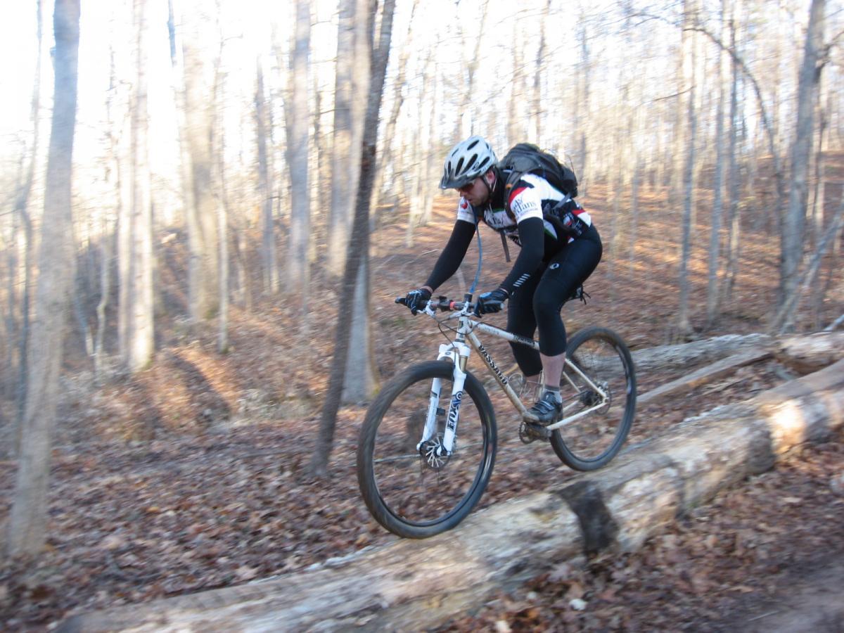 A mountain biker navigating over a fallen log on a forested trail, surrounded by autumn foliage and tall trees. The biker is wearing a helmet and cycling gear, actively balancing on the log as sunlight filters through the branches. Chicopee Woods mountain bike trail.