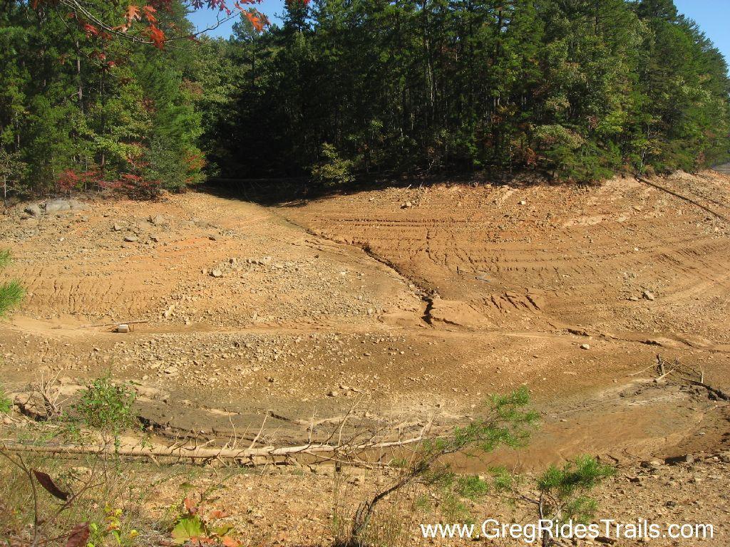 A dry lakebed with cracked earth and scattered rocks, bordered by lush green trees on one side, illustrating the impact of low water levels. Green Mountain mountain bike trail.