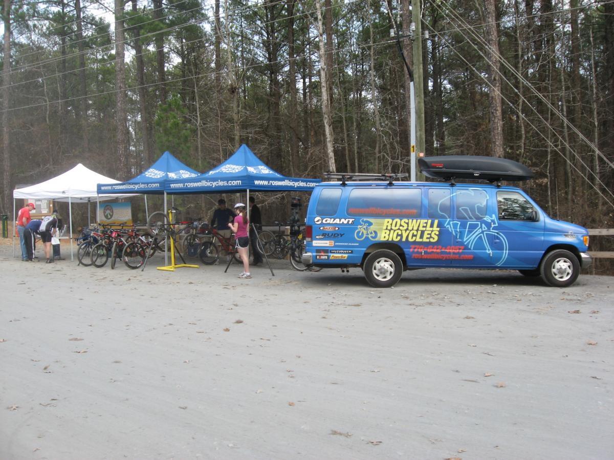 A bike shop setup featuring a blue van with "Roswell Bicycles" branding parked next to two canopies, one white and one blue, in a wooded area. Several bicycles are displayed in front of the tents, and individuals are interacting around the setup. The ground is a light dirt path with scattered leaves. Big Creek mountain bike trail.